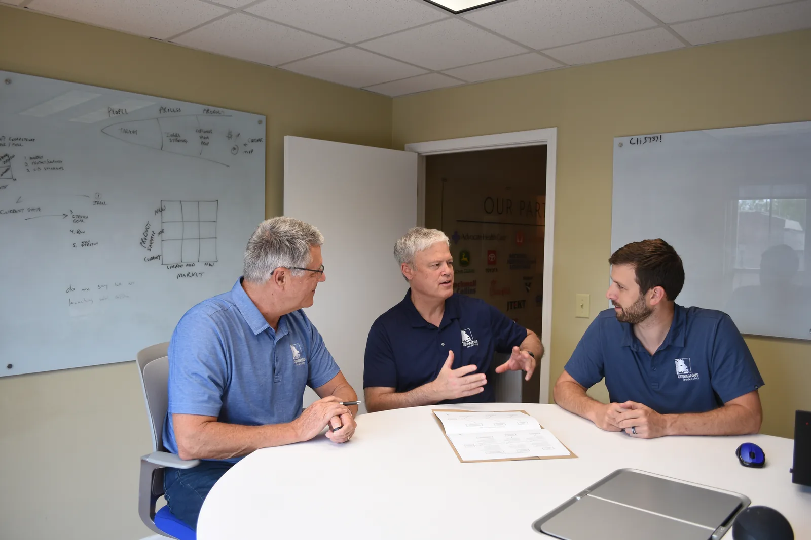 Three Courageous Leadership consultants in branded polos talking across a conference table, partner-logo wall in background